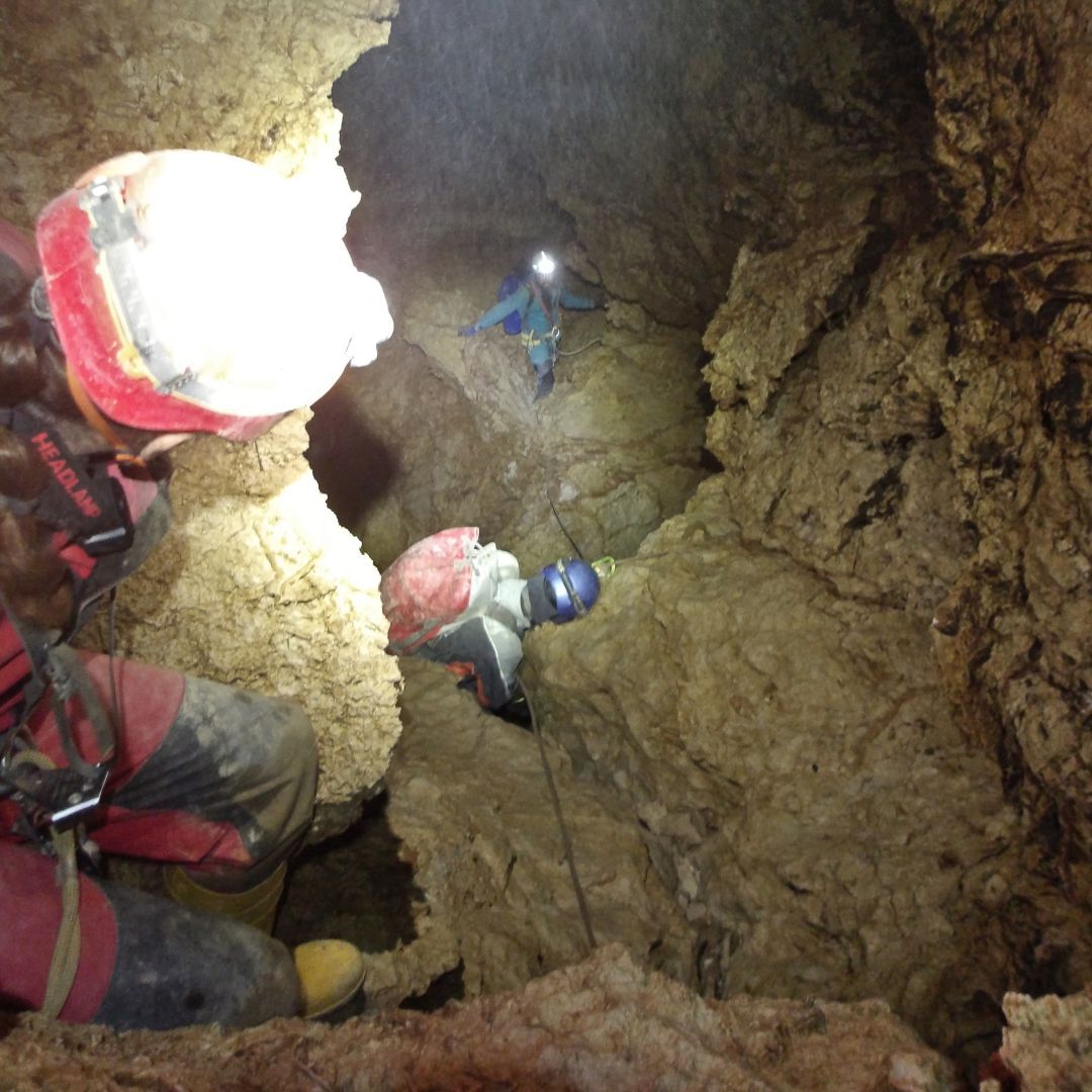 View down from over the shoulder of a person in caving suit with helmet and a duffle bag. Below are two more people in similar gear climbing down limestone rock with sharp edges and karst features, holding on to a rope. There's quite some spray illuminated in the headlamps.